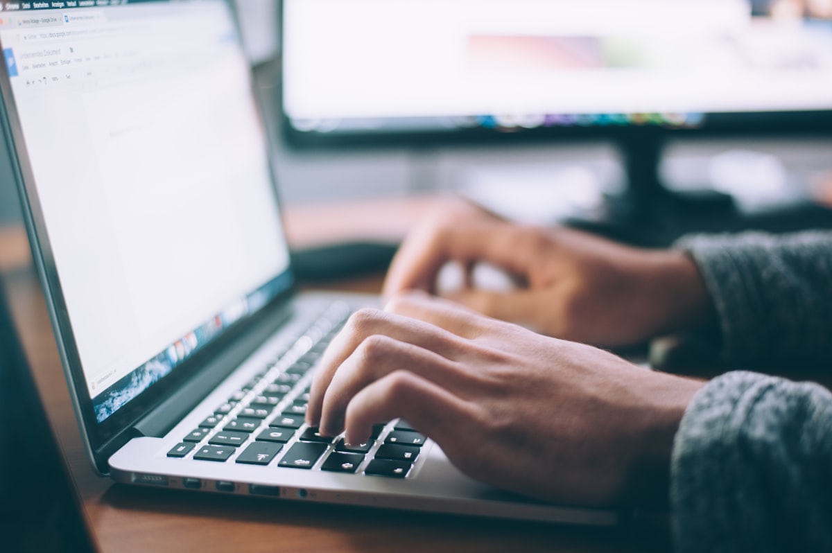 Person working on a laptop at a home desk in the evening with city lights visible through the window, representing side hustle work after a full-time job